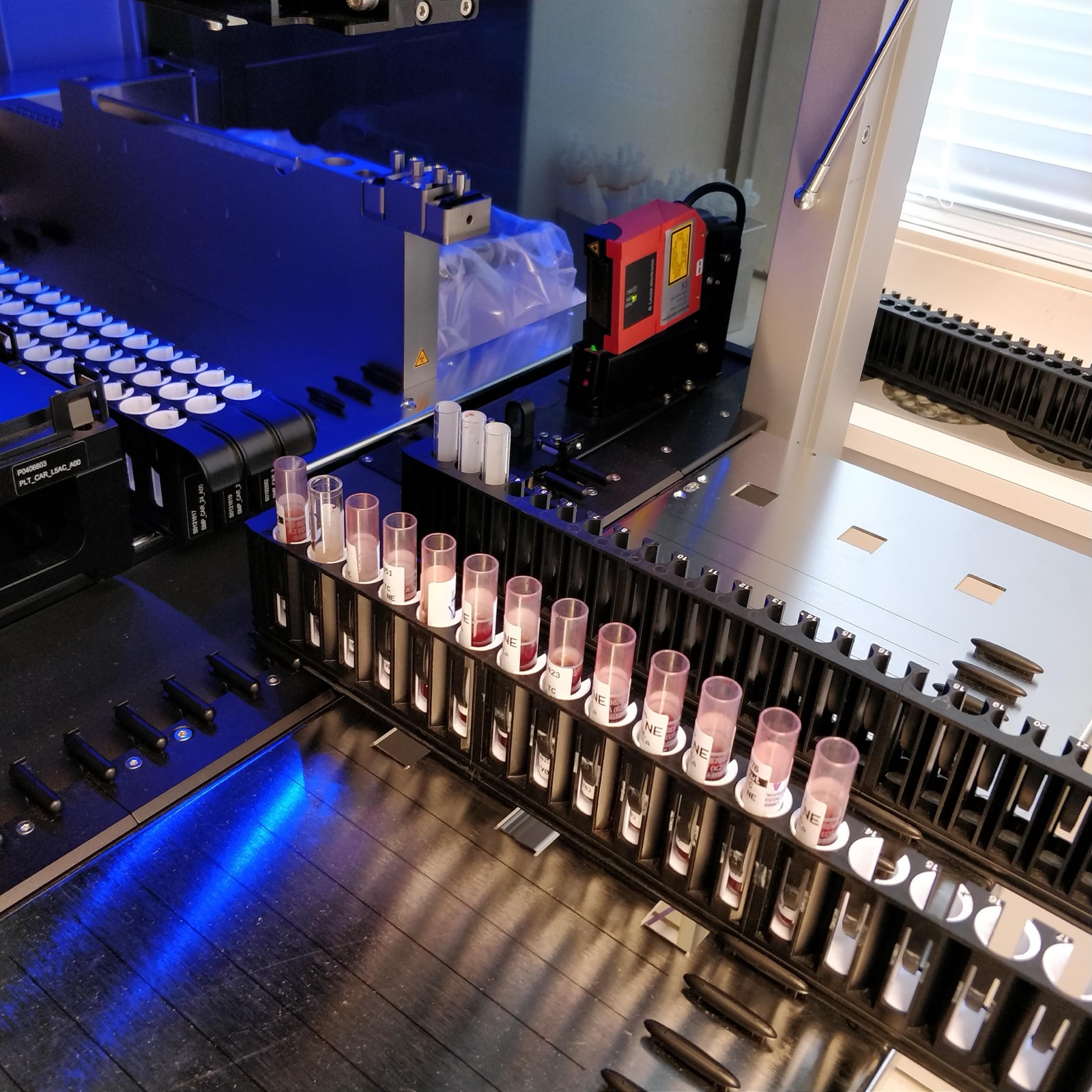 A Hamilton liquid handler deck with a red barcode reader mounted above the sample carriers, tubes standing in printed adapters along the carrier and blue instrument lighting on the deck behind