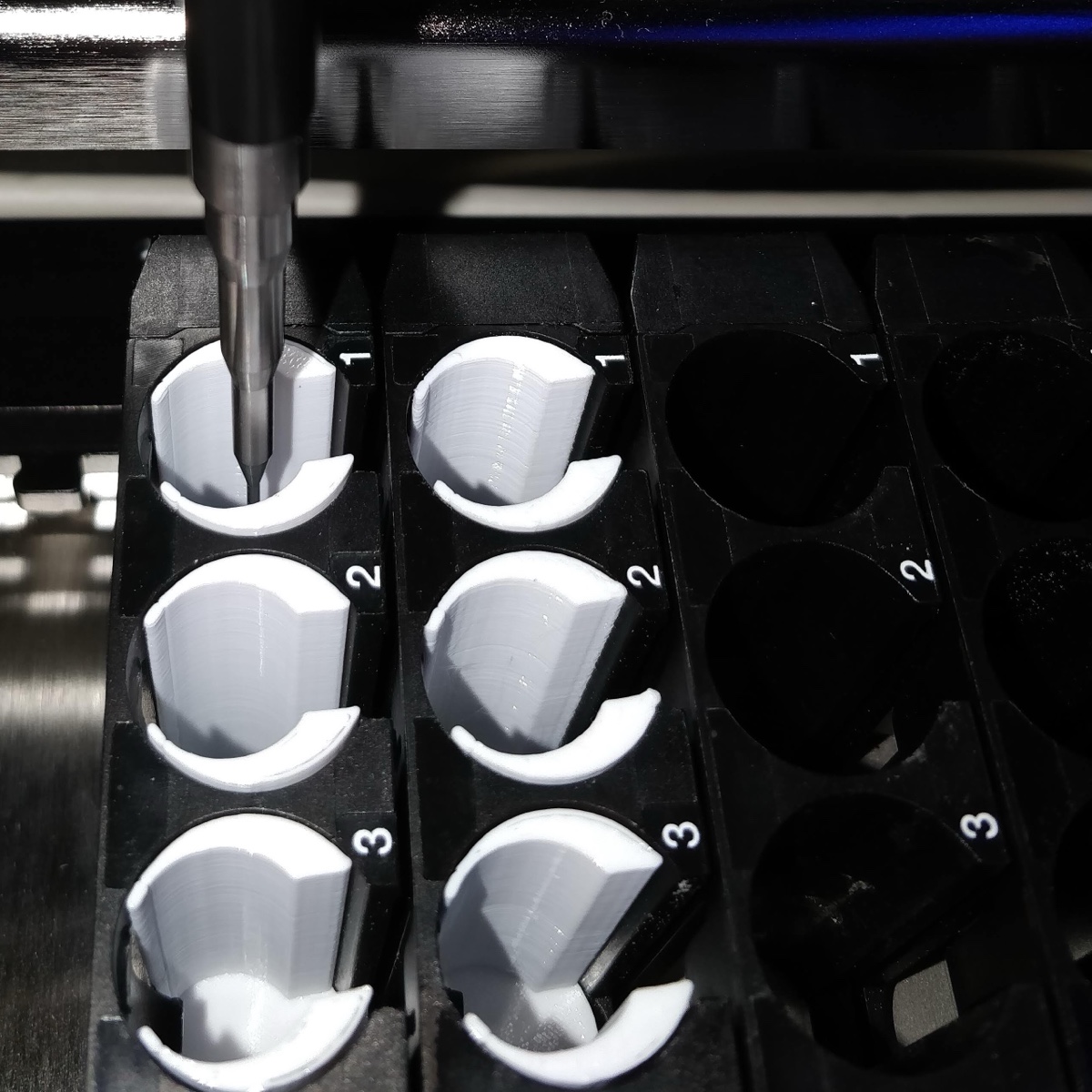 White 3D-printed tube adapters seated in the numbered positions of two black Hamilton sample carriers, a pipetting channel lowered into the first, with two bare carriers alongside