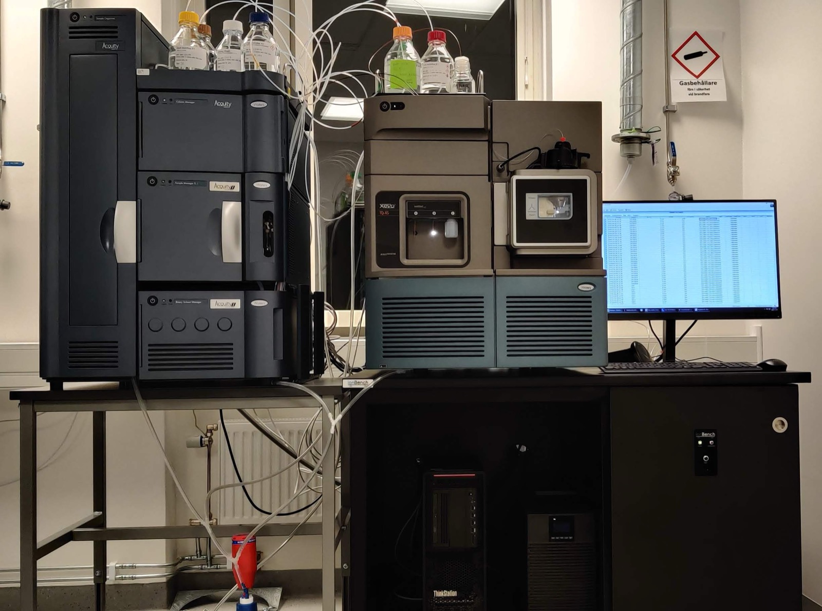 A laboratory LC–MS/MS bench: an ACQUITY UPLC sample organizer beside a stack of column manager, sample manager and binary solvent manager with solvent bottles standing on top, all on a metal table at left; a Waters Xevo TQ-XS and a workstation on a black IonBench at right