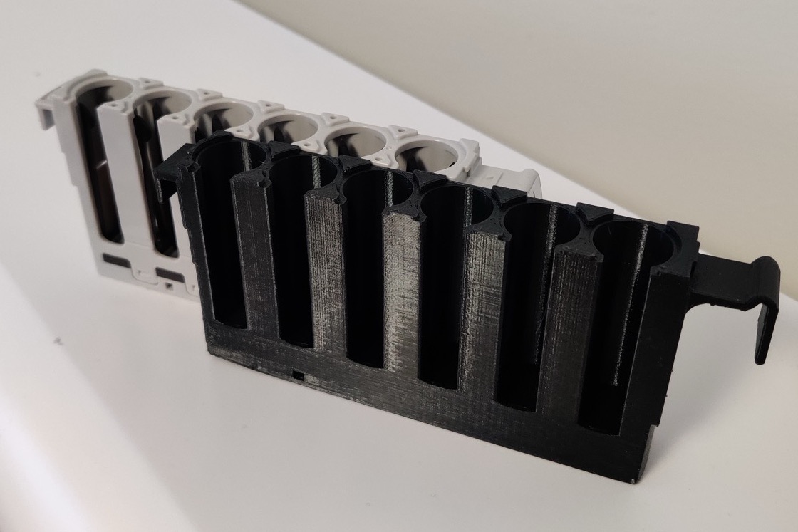 A black 3D-printed analyzer rack lying in front of the white OEM rack it copies, its wells cut deeper for taller collection tubes