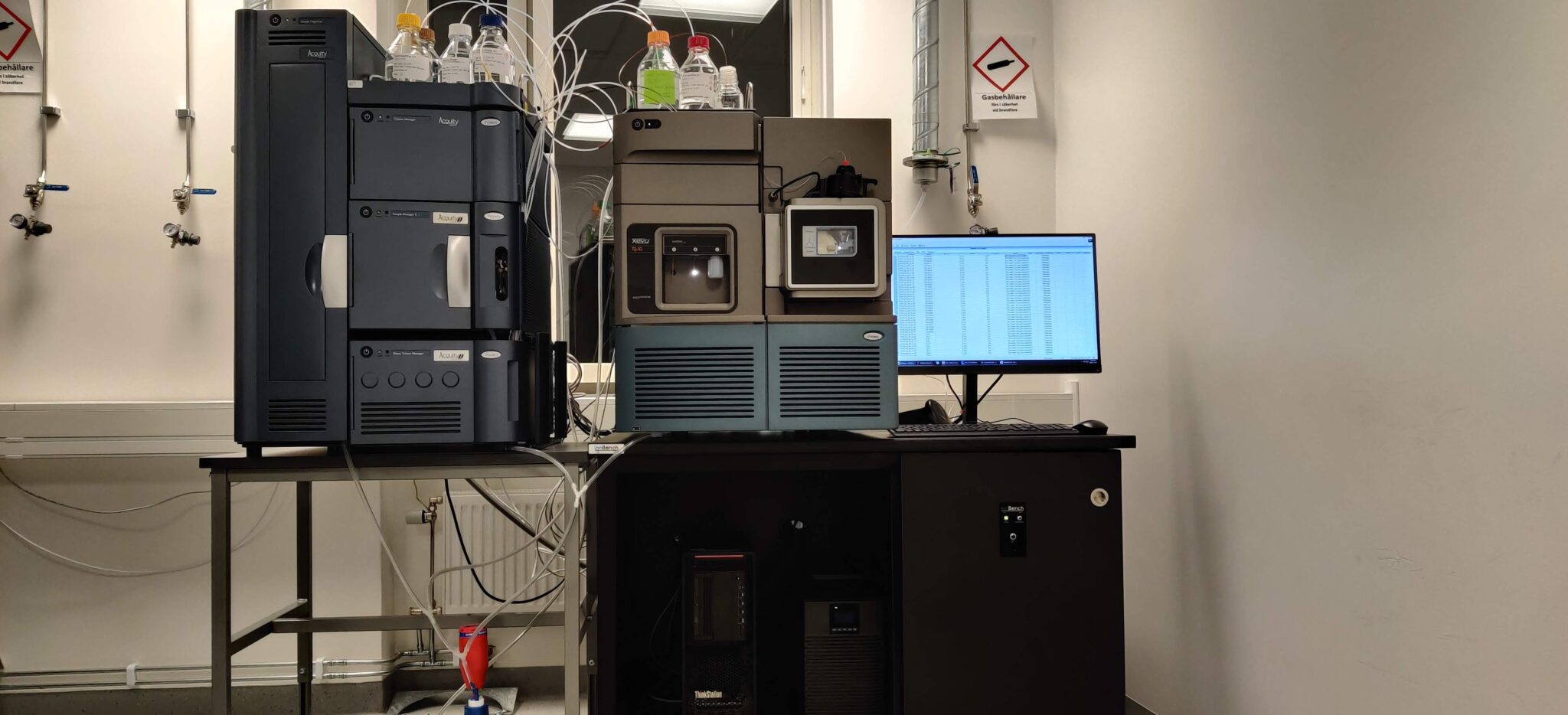 A Waters Xevo TQ-XS mass spectrometer and its ACQUITY UPLC stack on a bench during setup, solvent bottles on top, a workstation monitor beside it